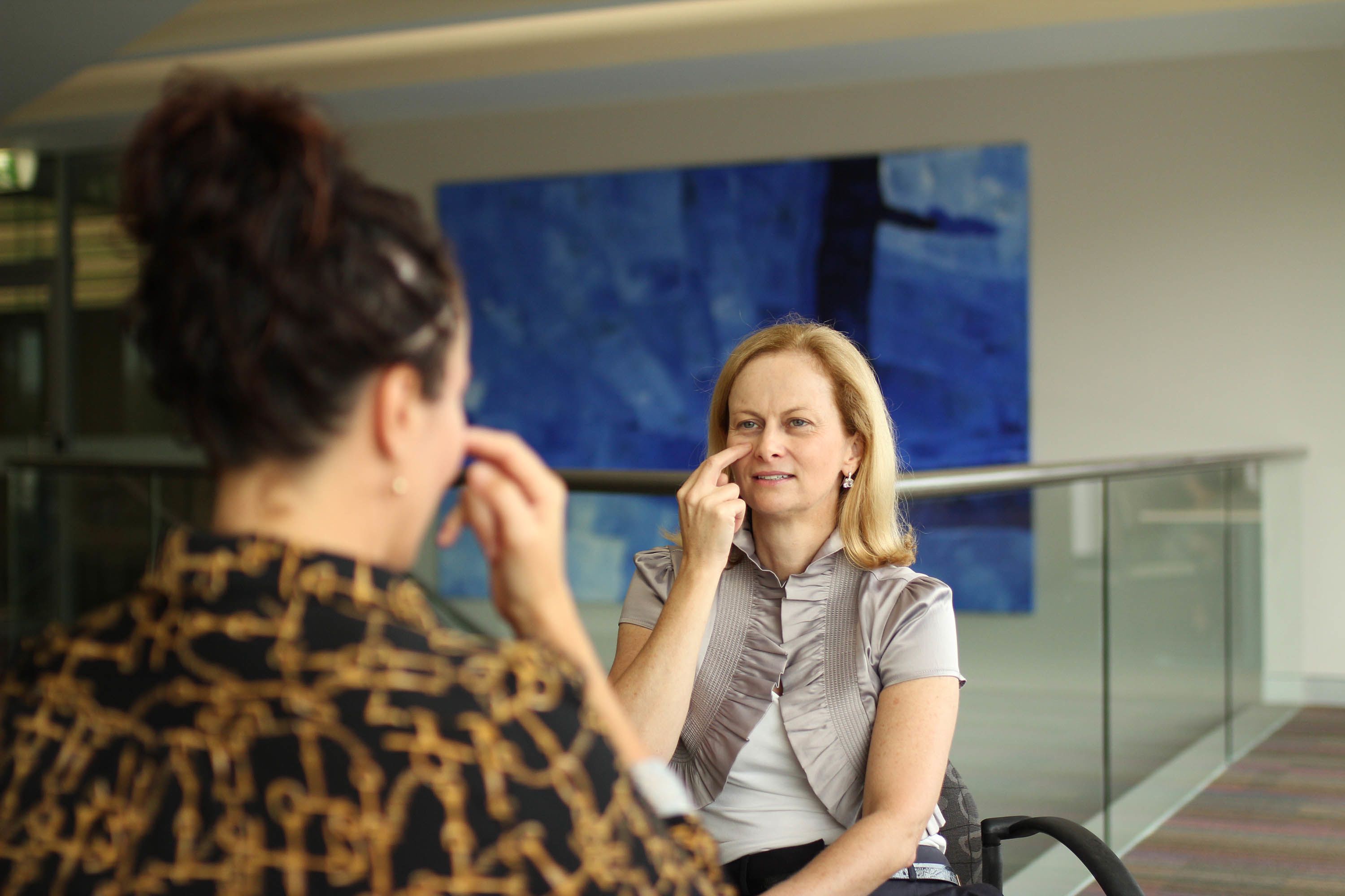 Two woman are facing each other, pressing two fingers to their face. 