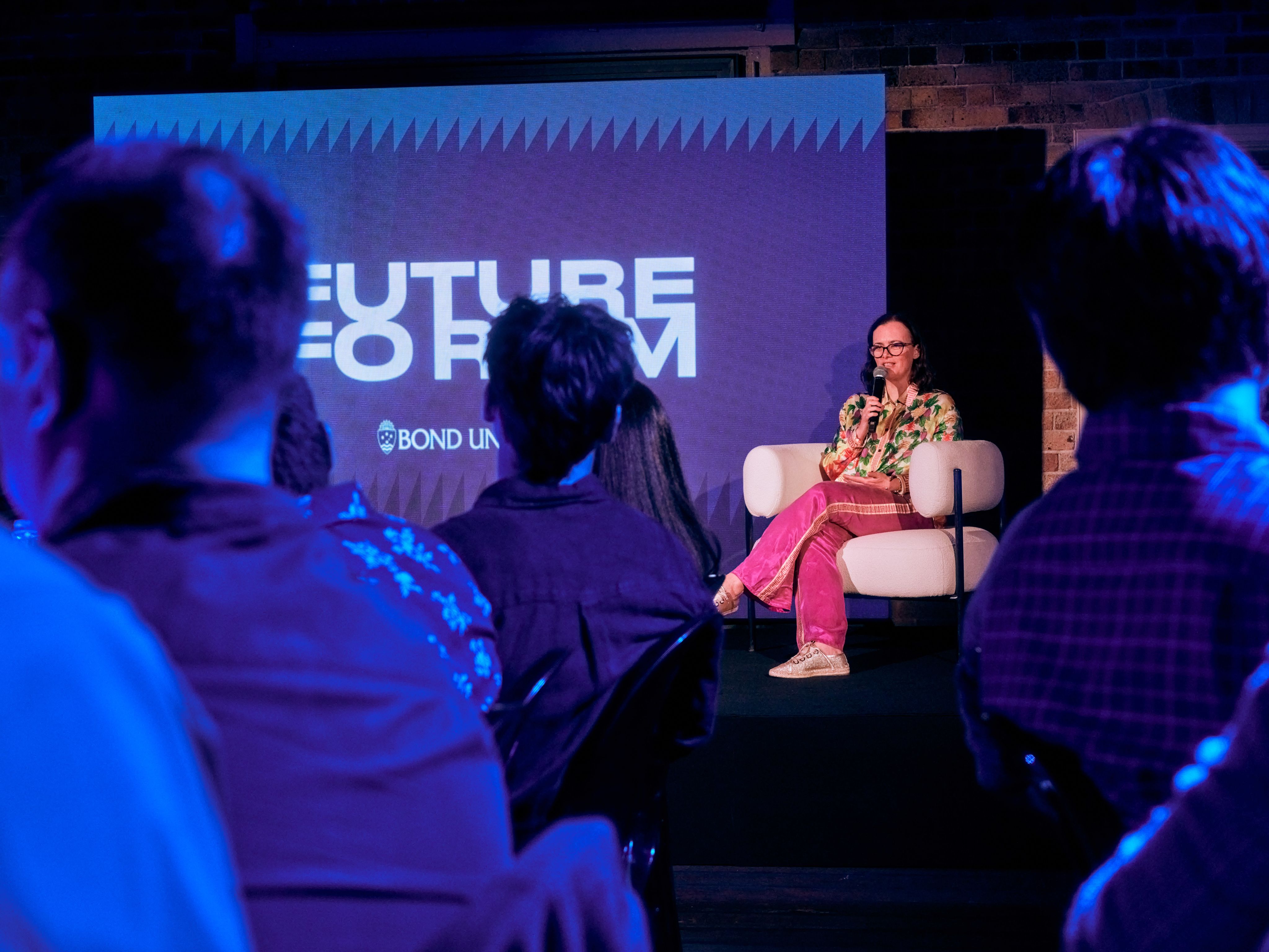 A woman is sat on a stage talking into a microphone.