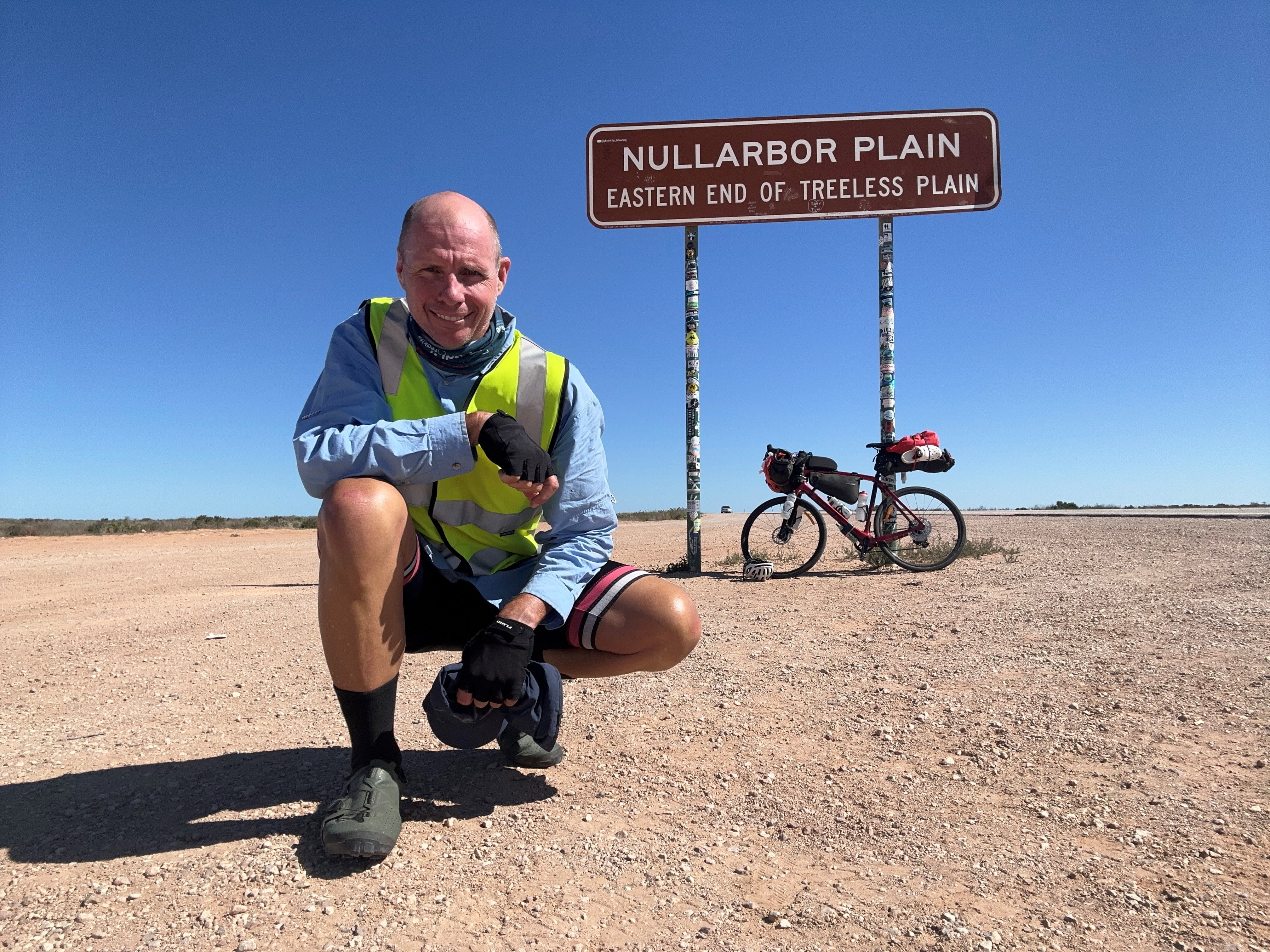 A man is crouched down on the dirt with a bicycle behind him,