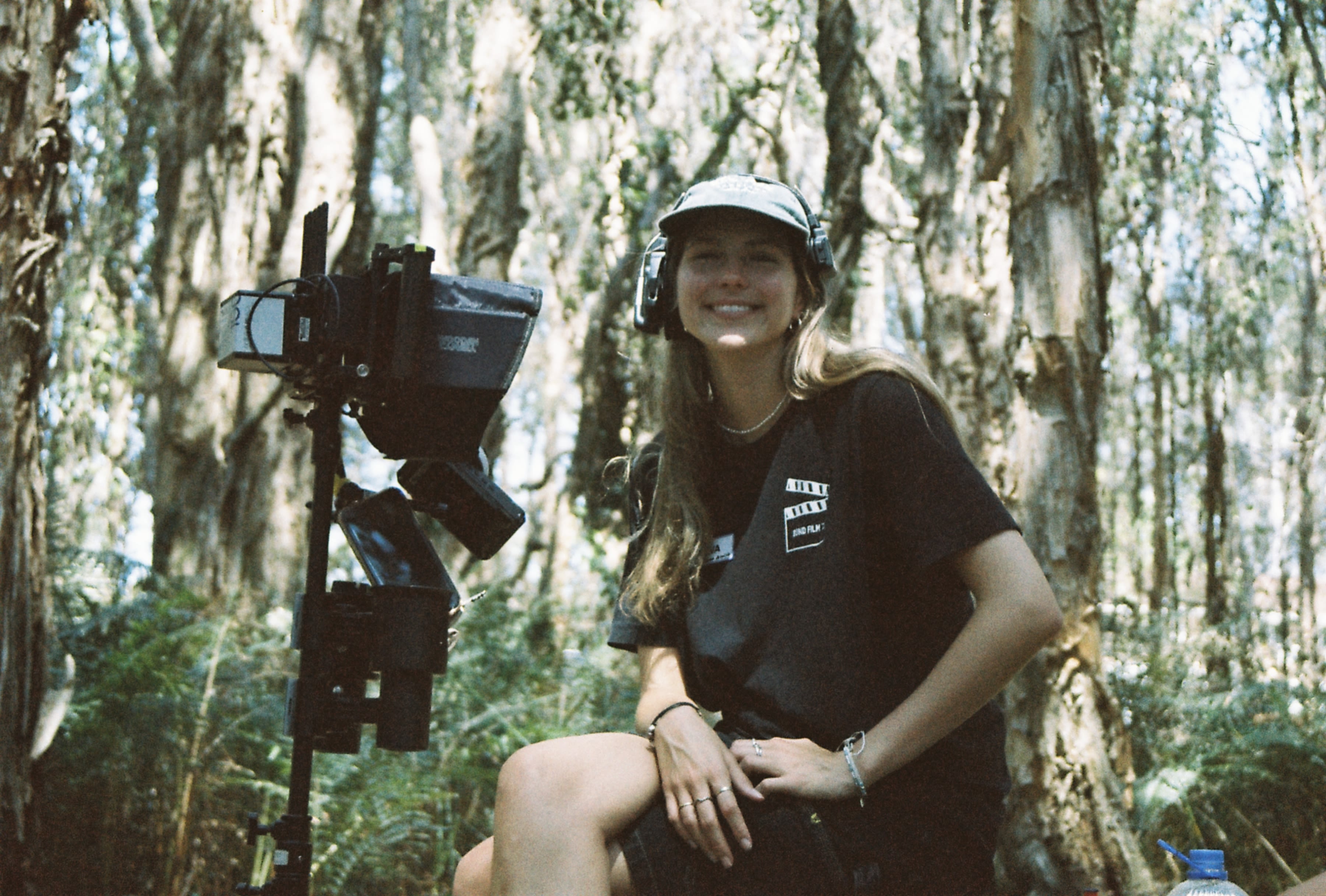 A woman wearing a hat and headset smiles in the forest, with a camera next to her