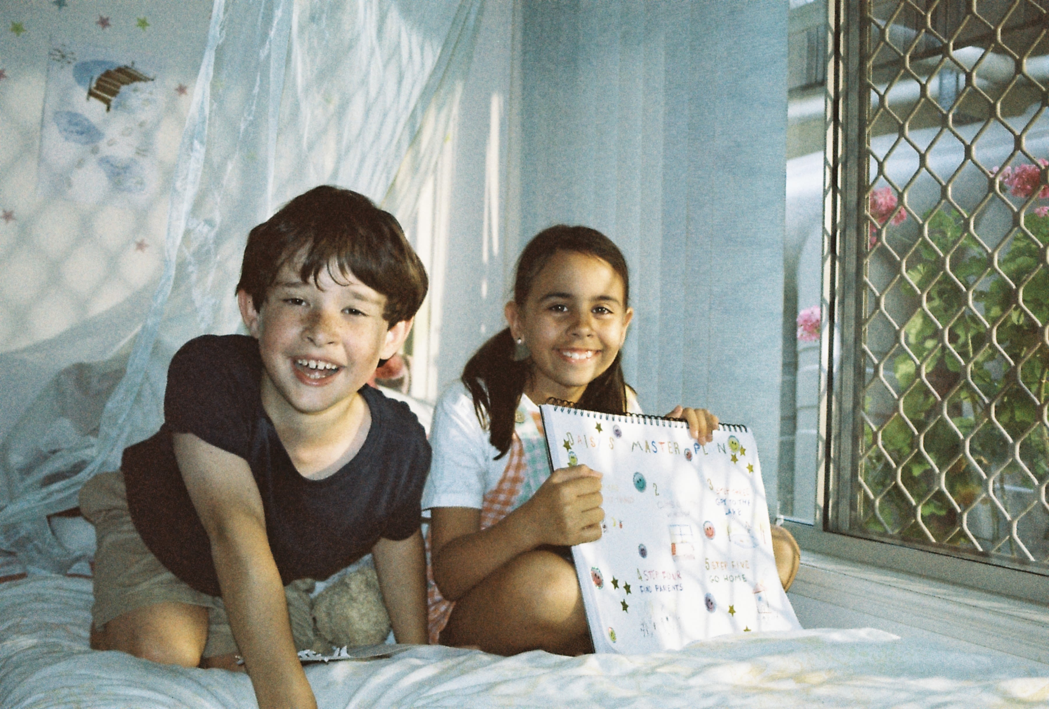 A young boy and girl sit smiling on a bed, the girl holding a map