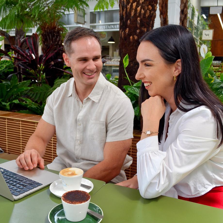 A man and a woman sit at a table looking at a laptop