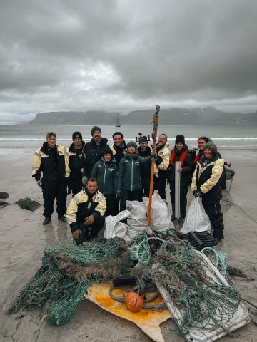 A group of people are standing on a beach with plastic debris. The sky behind them is grey. 