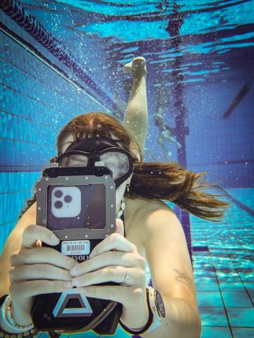 A young woman is underwater in a pool, holding out an iphone in a waterproof housing.