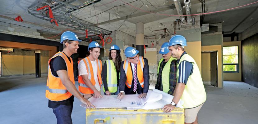 A group of people standing at a construction site, standing around a desk with paper on it.