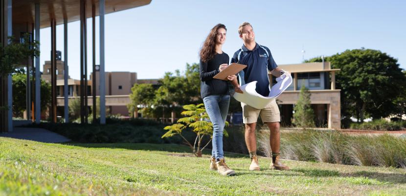 Two students standing on the grass in front of the Abedian School of Architecture building with a clipboard and paper.