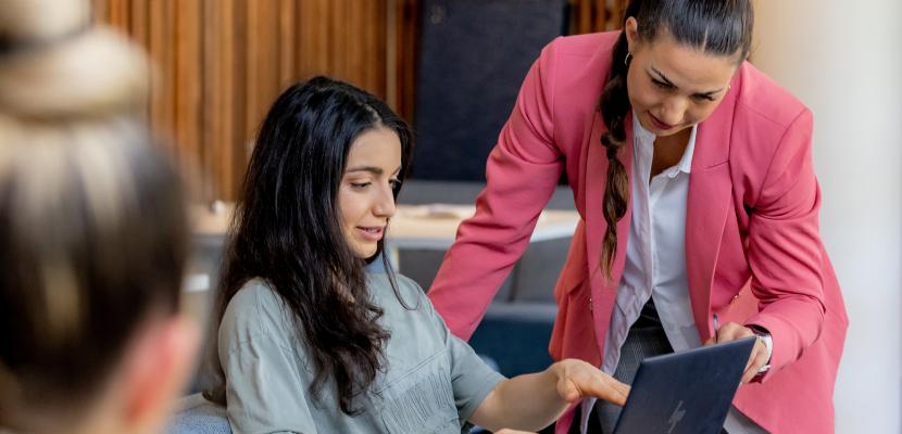 A woman in a pink blazer pointing at another woman's laptop screen.