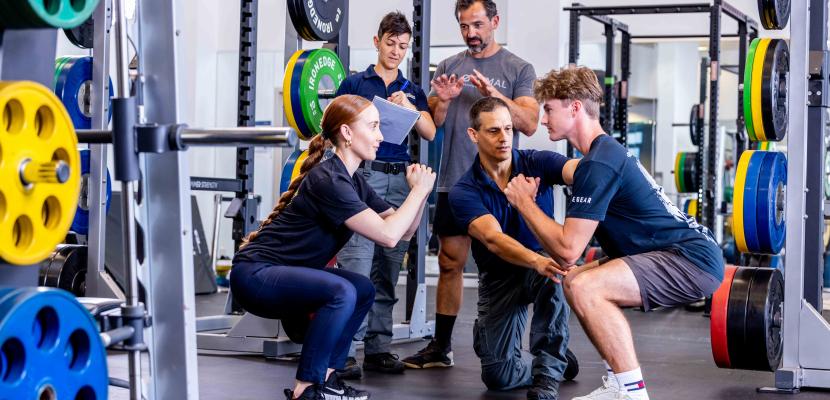 Two people squatting as three others watch and take notes of form in the Bond University Sports Centre.