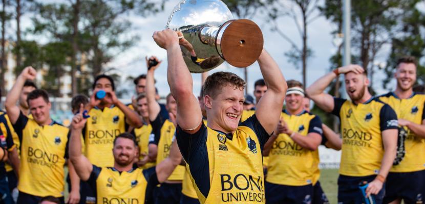 A member of the Men's Rugby Club holds a trophy above his head in celebration as his team celebrates behind him