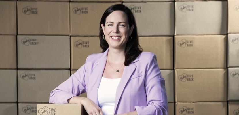 Carly Fradgley, a white woman in a pink blazer, stands and smiles with her hand resting on a Baby Give Back supply box