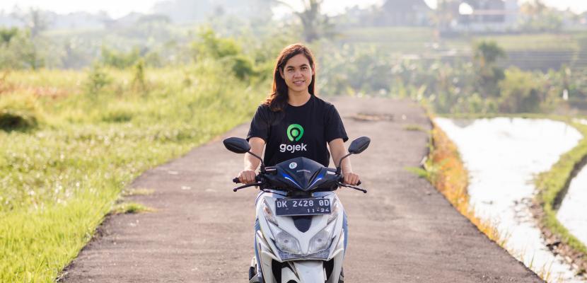 Bond alumna Tanah Sullivan is riding a Gojek motorbike wearing a Gojek t-shirt. She is riding on a dirt road through fields.