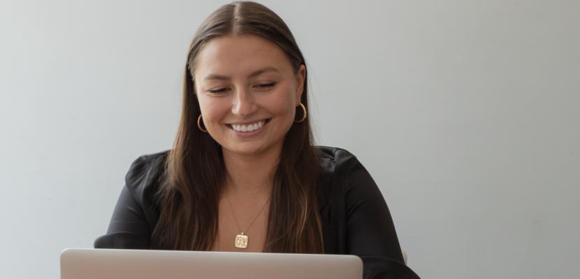 A woman with brown hair sits at a laptop smiling at the screen