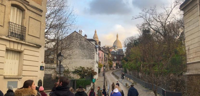 A group of people walk up a cobblestoned street with buildings either side. Sacre Coeur is visible in the background.