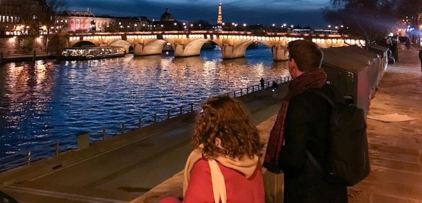 Two people stand facing the River Seine at night time. Everything is lit up.