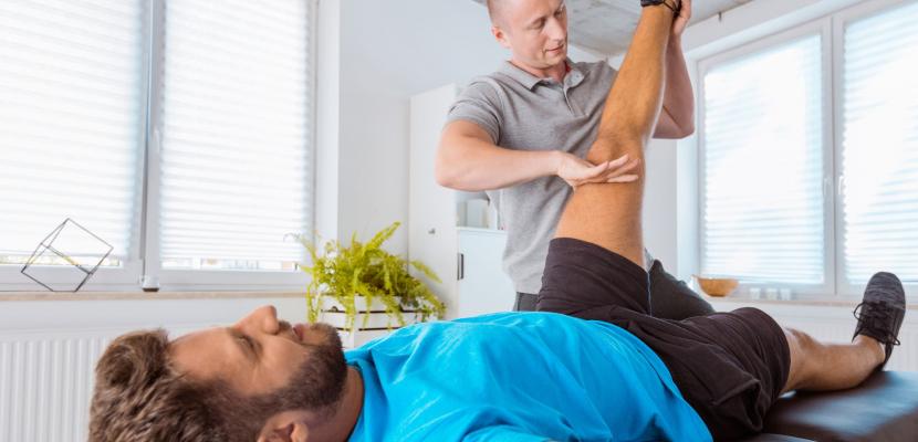 A patient lies on a bed and the phsyiotherapist is lifting his leg into the air