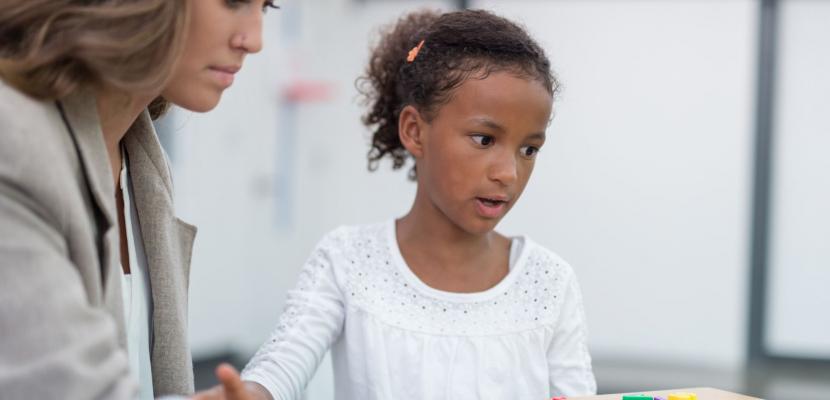 A woman sits with a child at a desk, they have coloured number and letter blocks on the table