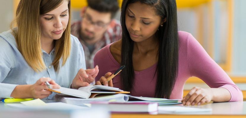 Two women sit at a desk, in a classroom, reading a textbook