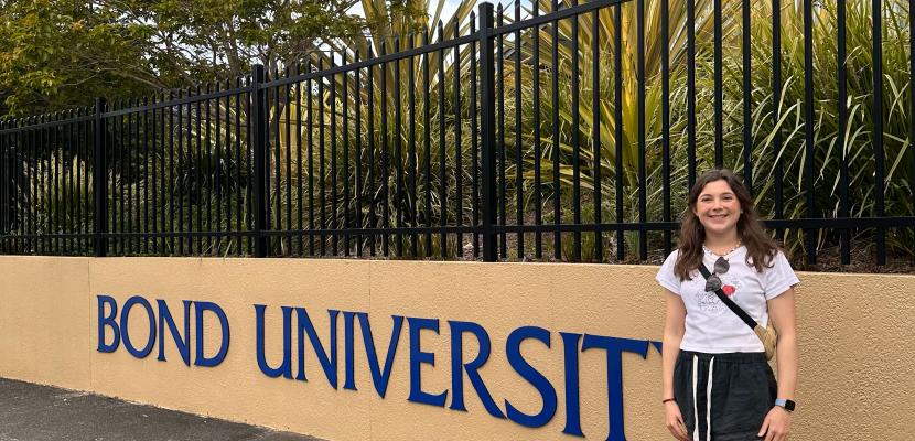 A girl with brown hair stands next to a sign that reads Bond University, smiling