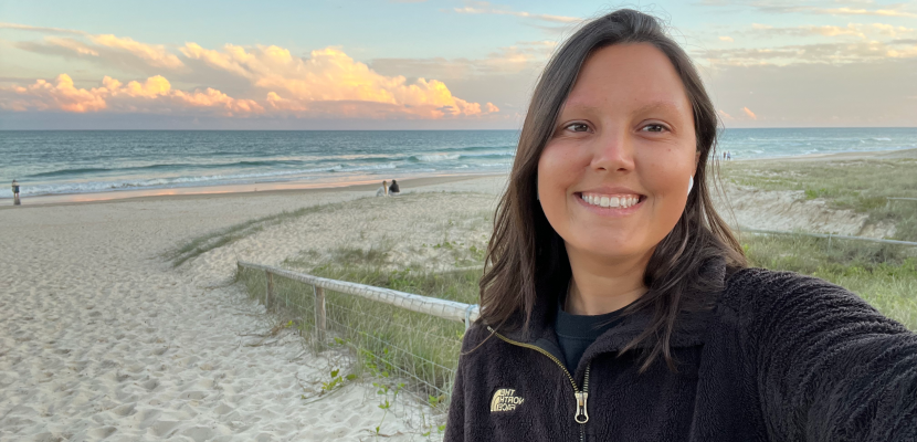 Maju, a woman with brown hair and tan skin, is smiling with the beach and a beautiful sunset behind her