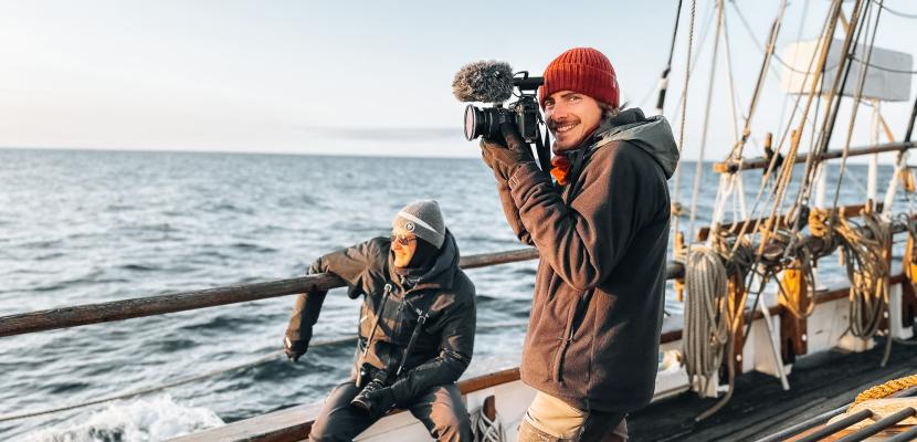 A man is holding a video camera upon on a sailboat. 