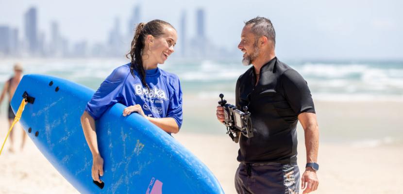 A man in a black rash shirt is holding filming equipment on a beach talking to a young woman holding a surfboard.