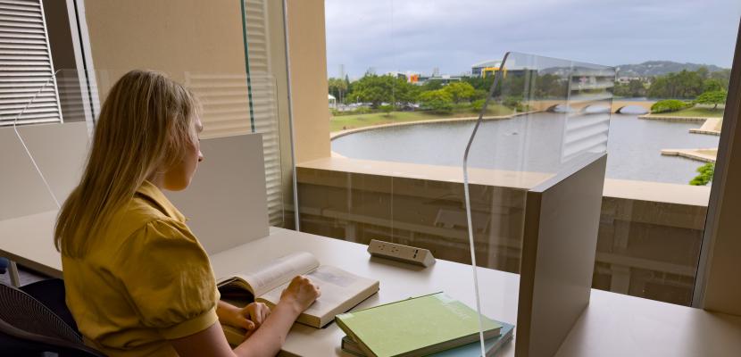 A young woman in a yellow dress is sat at a desk studying with a view of a lake in front of her.