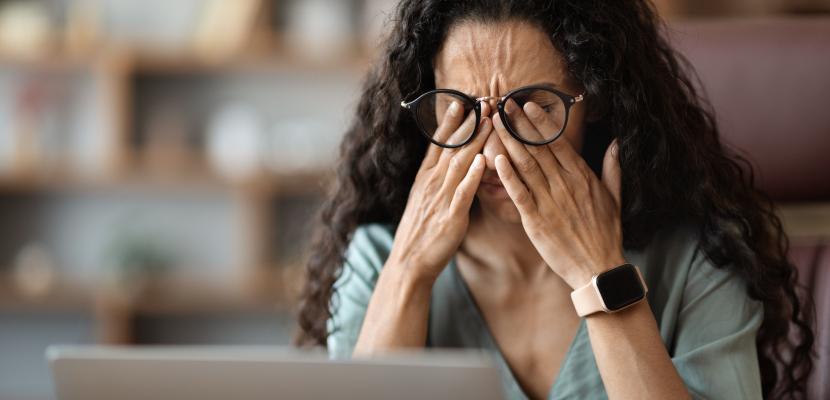 Woman with long dark curly hair and glasses rubbing her eyes in front of a laptop