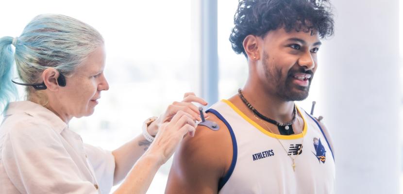 A person in a sports jersey has a wearable sensor being attached to the upper arm by another person in a bright indoor setting.