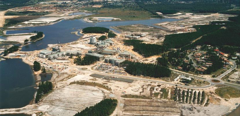 Aerial photograph of a construction site beside a lake with remnant pine forests and a small residential area.