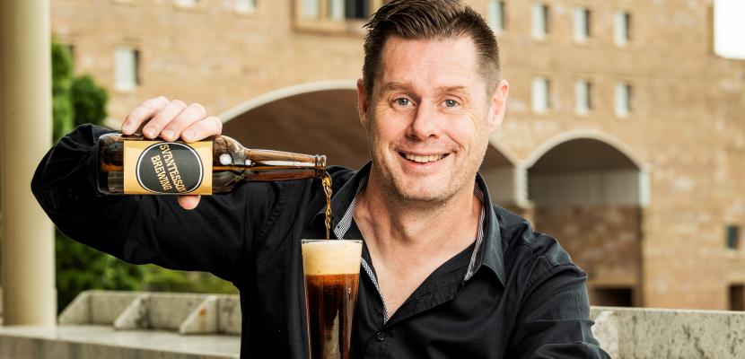 A man with short brown hair smiles at the camera as he is pouring a dark beer into a glass