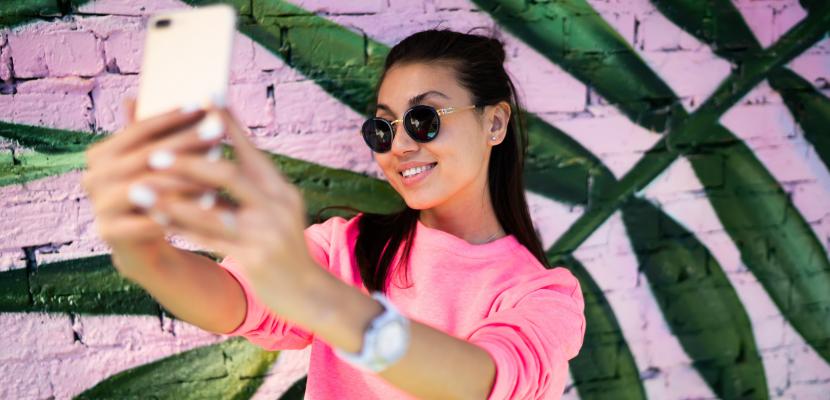 A girl in sunglasses smiling while taking a selfie with her mobile phone