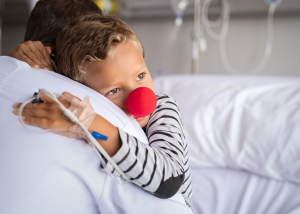 A young child is sitting in a hospital bed wearing a red clip on nose and hugging an adult