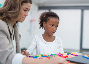 A woman sits with a child at a desk, they have coloured number and letter blocks on the table